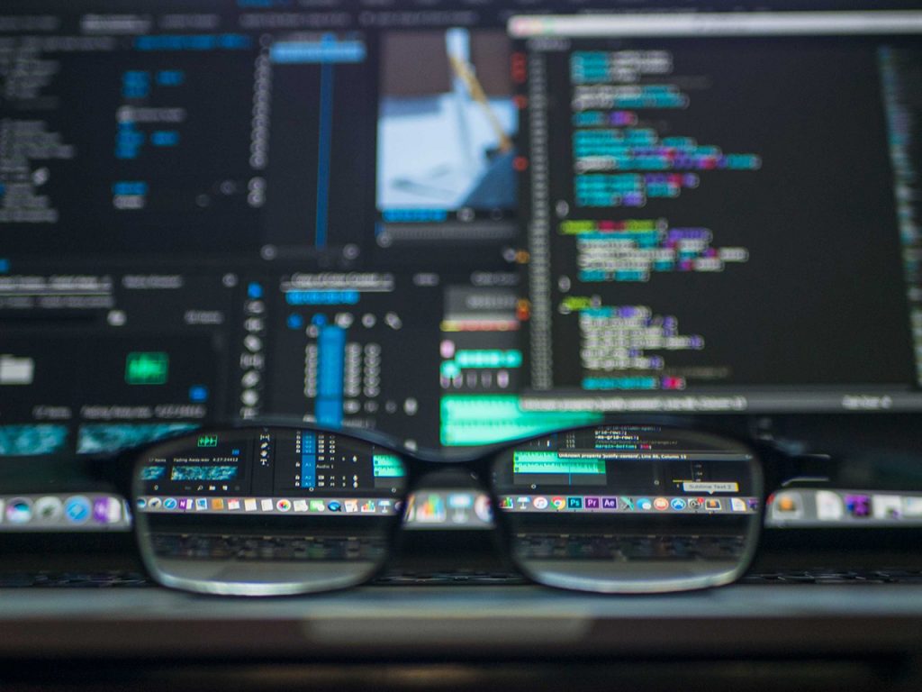Glasses on a computer desk with a busy code screen in the background
