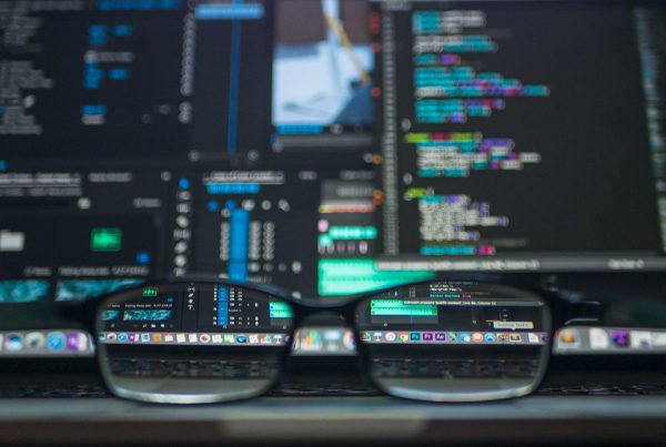 Glasses on a computer desk with a busy code screen in the background