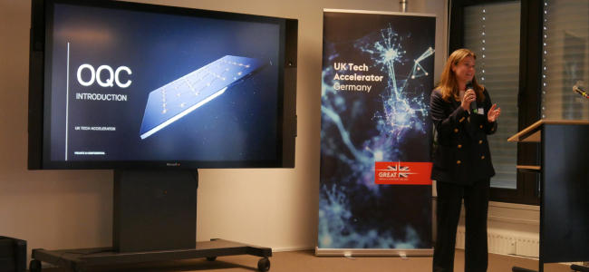 OQC's Head of Commercial, Caro Ehrman, on stage with a microphone speaking. Behind Caro is a large screen with a graphic of an OQC quantum chip, with overlaid text which reads 'OQC, Introduction, UK Tech Accelerator, Private and Confidential' and an exhibition pop up banner which reads, 'UK tech Accelerator Germany.