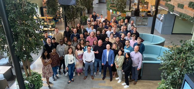 Around 70 OQC UK team members standing as a group in a wide corridor, all facing the camera for a 'team photo