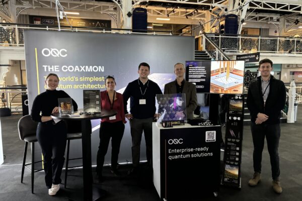 OQC booth background consisting of a black backdrop with OQC logo and chip render. Tall table with 2 stools, TV screen and brochure display and table. Five OQC employees standing in the booth posing for a photo.