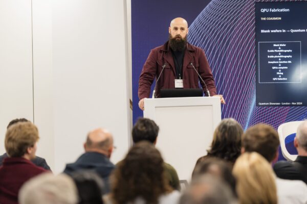 Image of male presenter standing behind a podium facing the audience. Screen behind speaker displaying information regarding 'QPU Fabrication'. Blurred backs of audiences heads in foreground of photo.